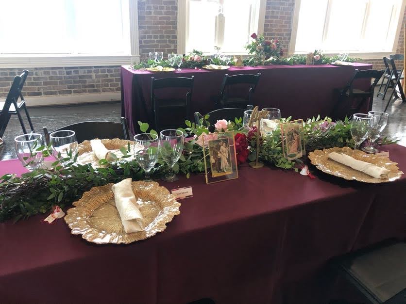 Decorated banquet tables with burgundy tablecloths, gold charger plates, folded napkins, wine glasses, and greenery garlands, featuring small framed photos, arranged in a bright brick-walled venue with large windows.