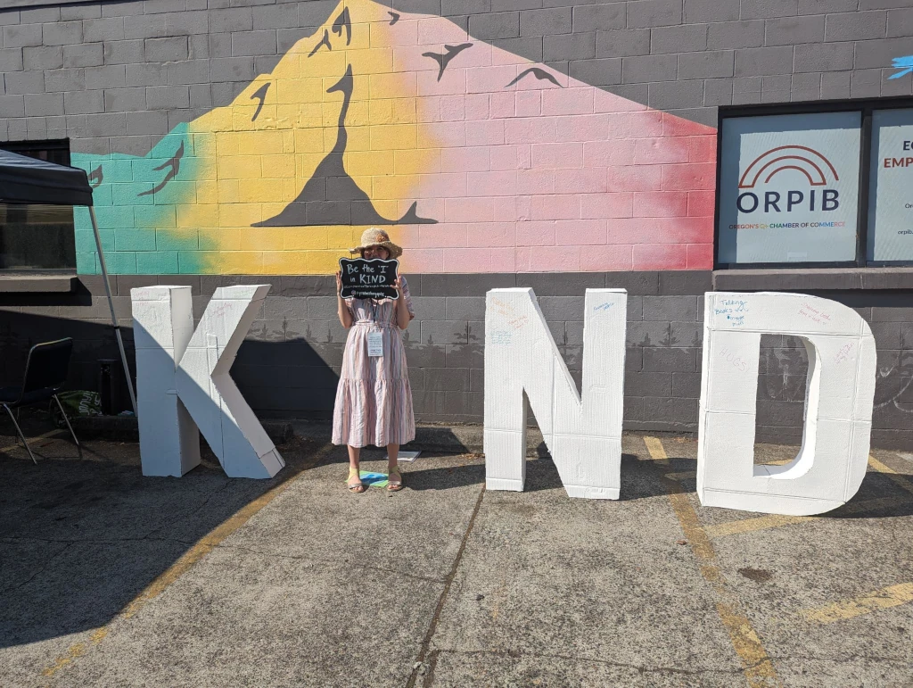 Amanda Sparks standing between large white letters spelling KIND, holding a chalkboard sign that reads "Be the I in Kind," in front of a colorful mountain mural at an outdoor event.