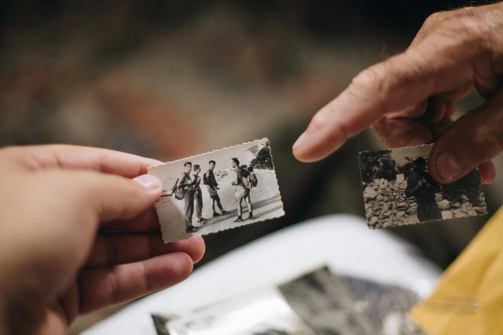 Two people sharing old black-and-white photographs, one person pointing to a photo showing a group of young men outdoors