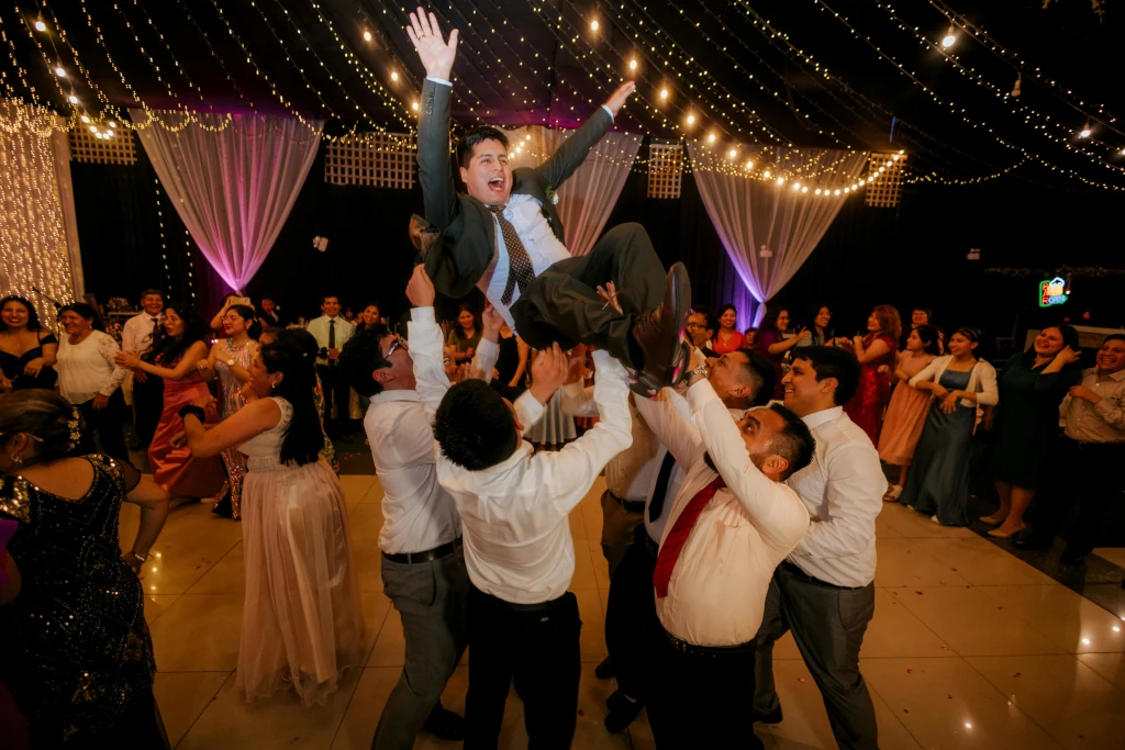 Wedding guests lifting a celebrating man into the air on a packed dance floor, surrounded by string lights and a full crowd of reception guests