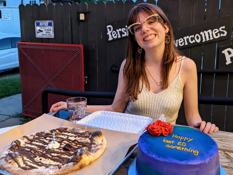 A smiling young woman at an outdoor table with a birthday cake iced "Happy last 20 something."