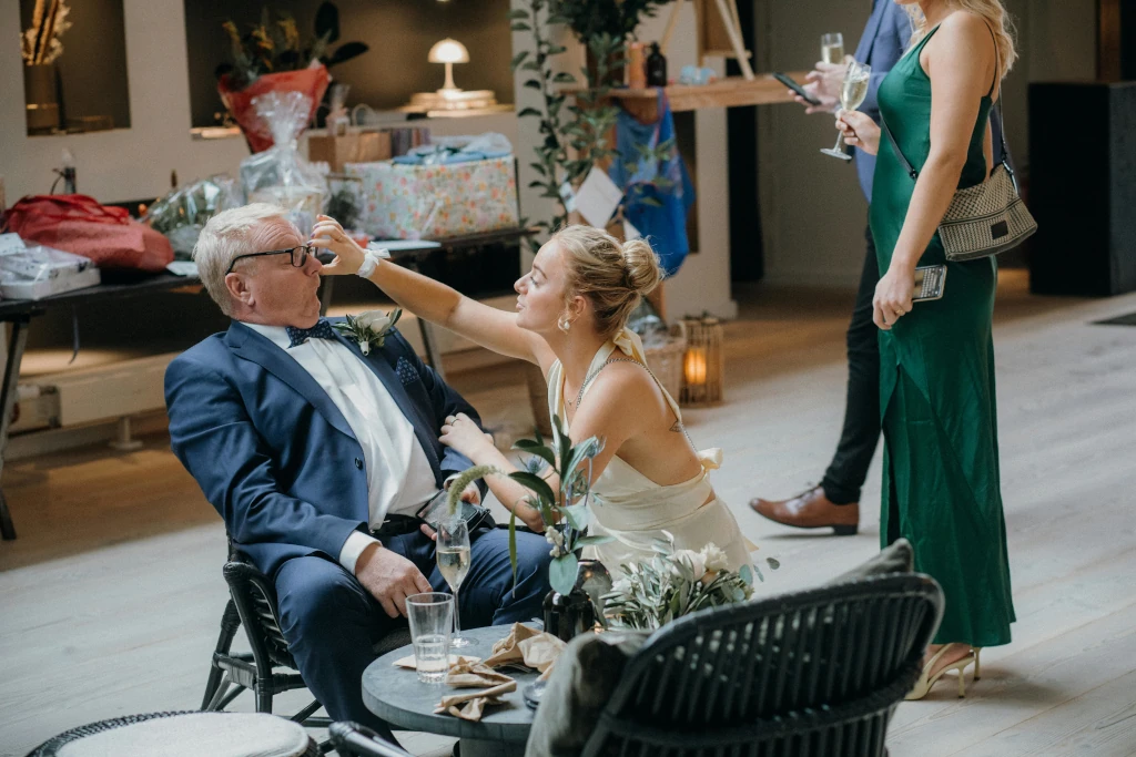 Woman kneels beside an older man at a reception and playfully takes his glasses as guests and gifts appear in the background.