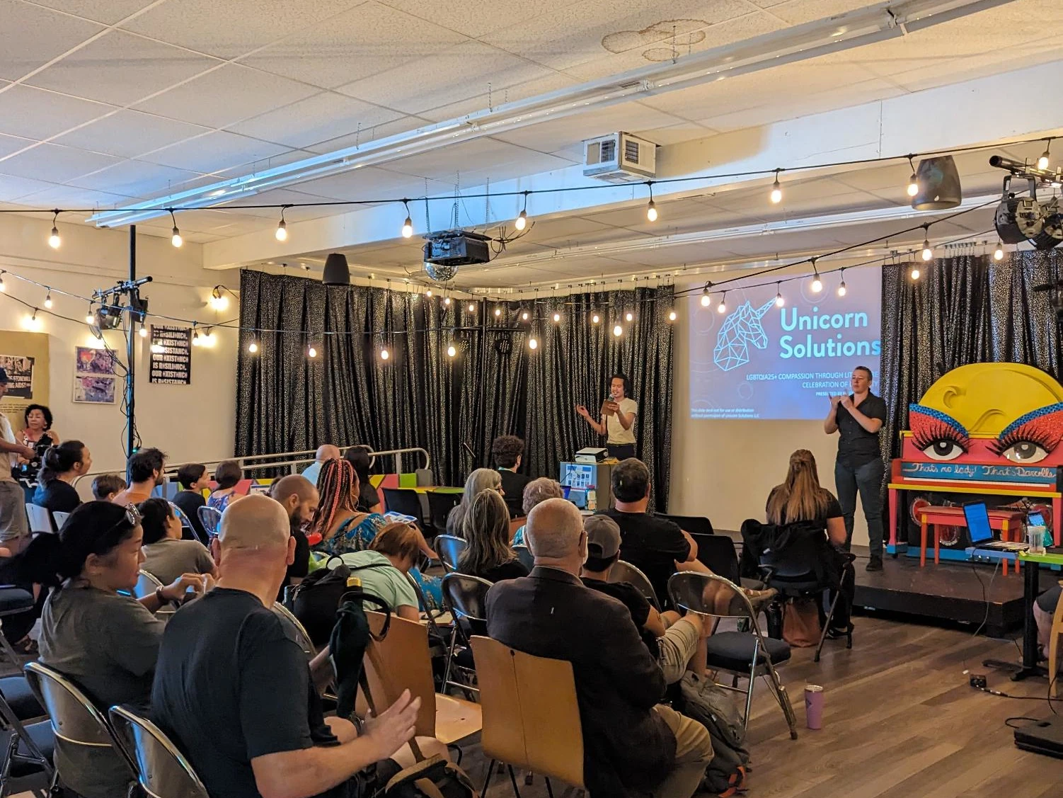 A packed community event at the Q Center in Portland, with an audience seated facing a presenter on stage next to a projected "Unicorn Solutions" screen. String lights and sparkly black curtains hang above the stage, with colorful murals and a painted piano visible on the right.