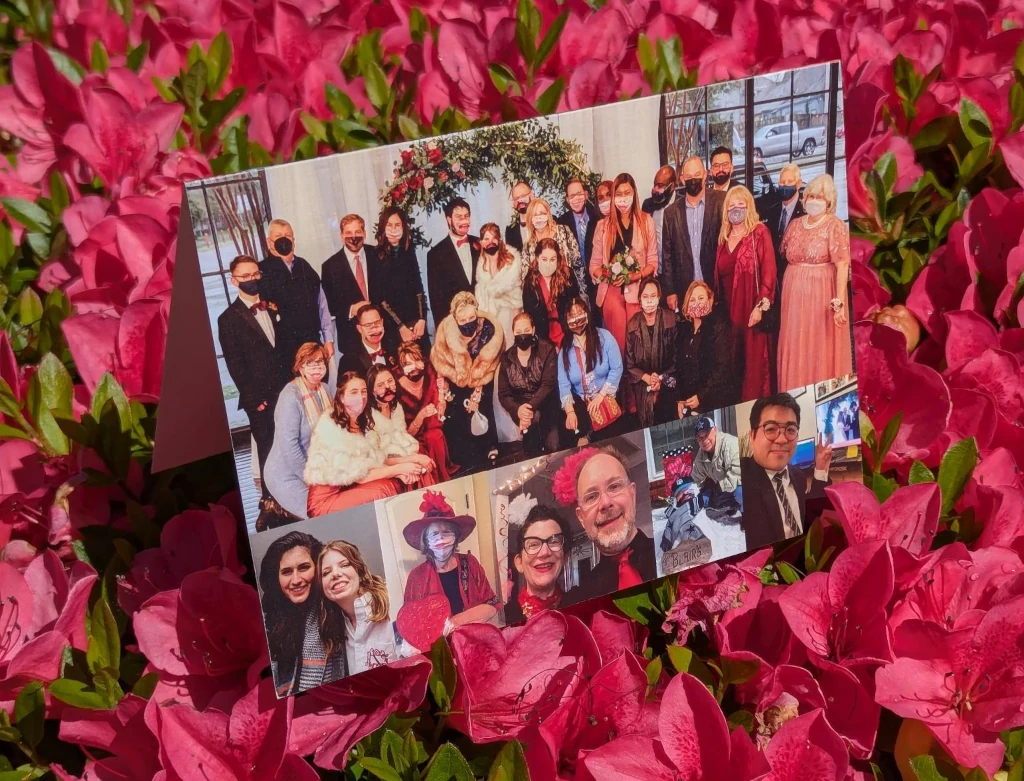 A folded photo card stands among vibrant pink flowers. The card features a large group photo of people dressed formally at an indoor celebration, many wearing face masks, along with several smaller candid portraits arranged beneath it. The bright floral background and collage-style images give the scene a warm, commemorative feel.