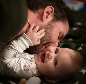Close-up of a man kissing a smiling baby on the cheek, both gazing lovingly toward the logo in the center.