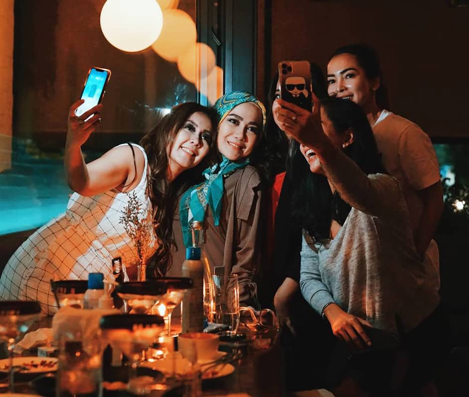 Five women gathered around a dinner table, smiling and taking selfies together in a warmly lit setting.